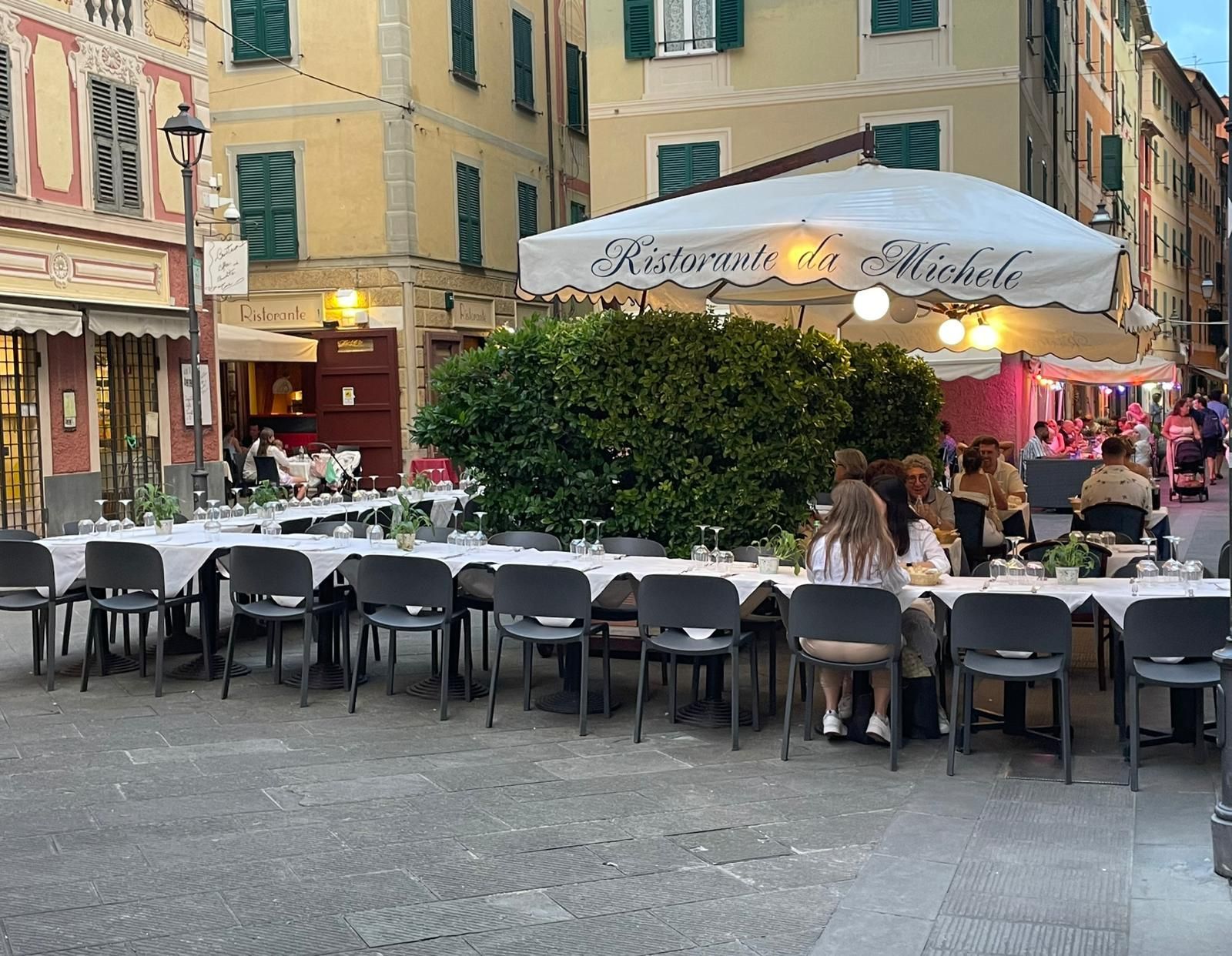 Il Ristorante da Michele offre una zona pranzo all'aperto in una piazza italiana, con tavolini all'ombra di ombrelloni e un grande cespuglio.