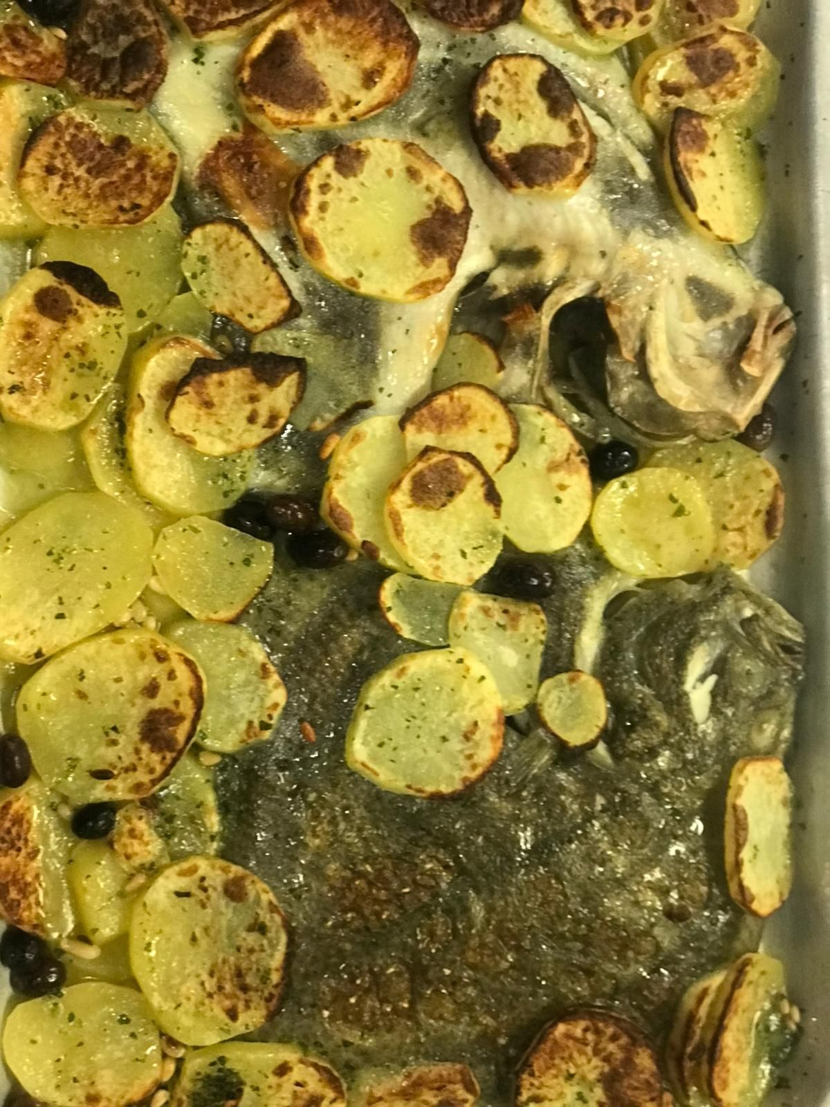 Un pesce piatto intero arrostito, probabilmente rombo o sogliola, disposto in un vassoio di metallo con patate a fette dorate e olive.