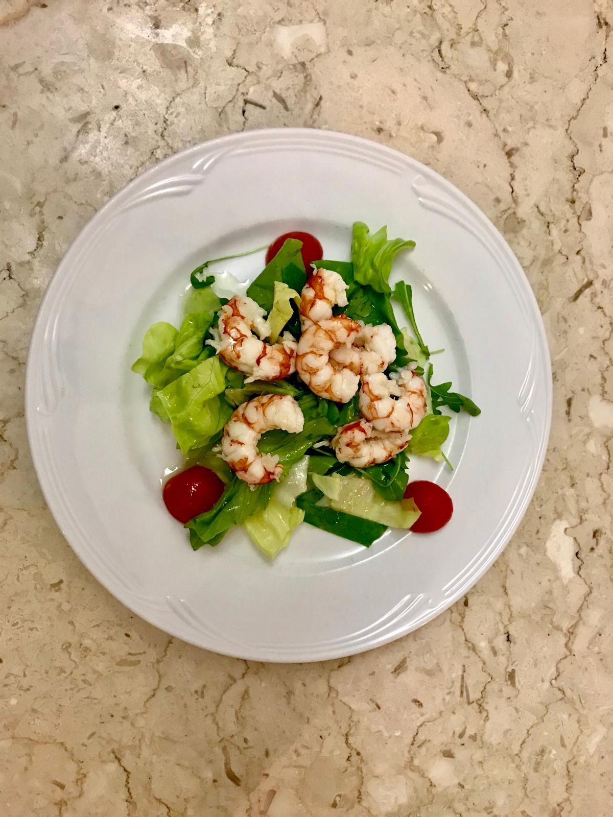Una piccola insalata di gamberetti con lattuga e pomodorini, servita su un piatto bianco su un piano di lavoro in marmo.