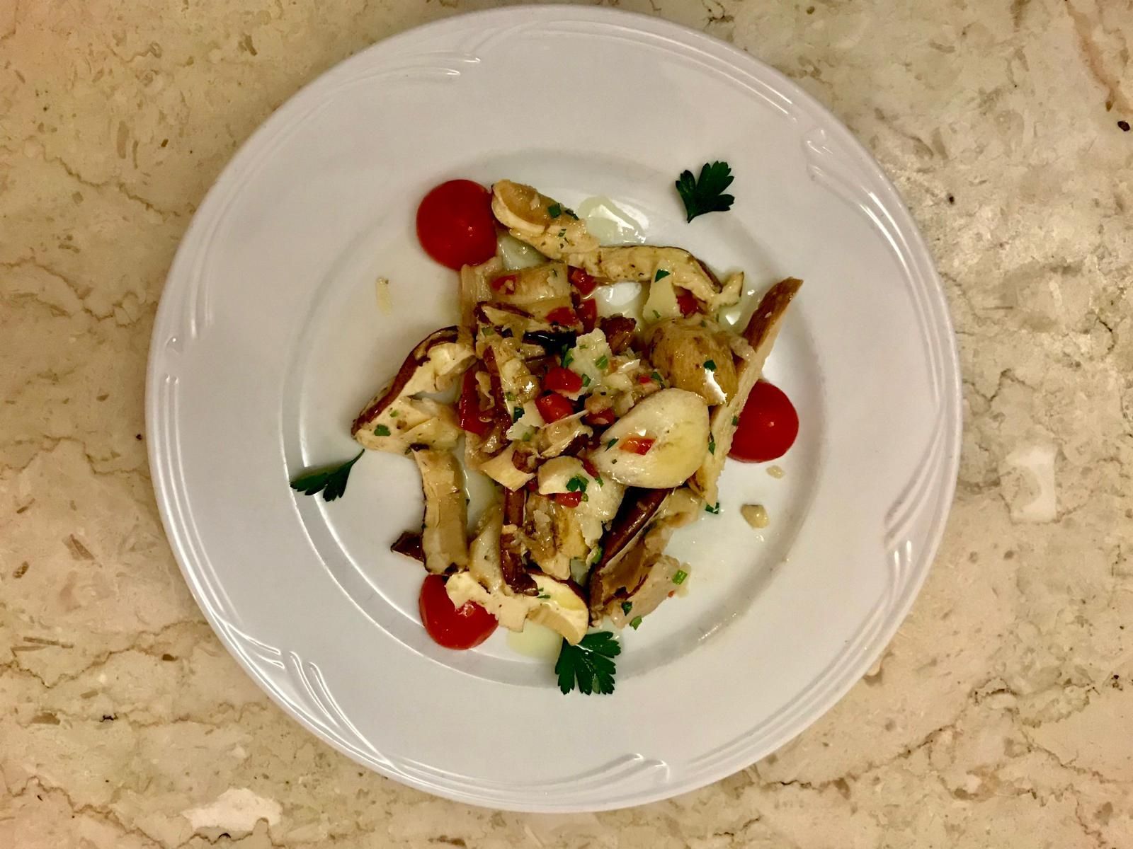 Un piatto bianco su una superficie di marmo beige, contenente funghi affettati, pomodorini e prezzemolo fresco.