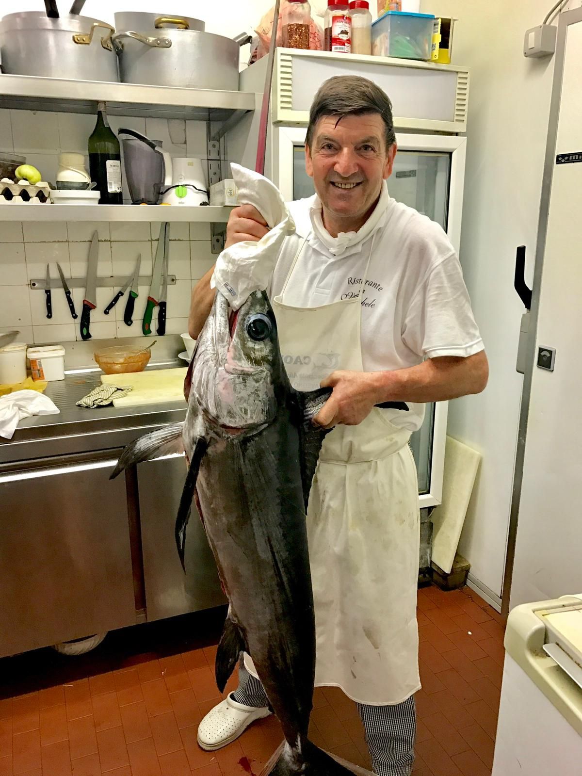 Uno chef in uniforme bianca tiene in mano un grosso pesce intero all'interno di una cucina professionale.