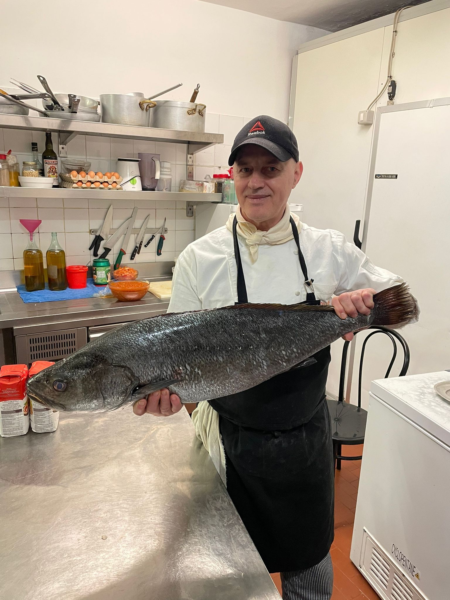 In cucina, uno chef in uniforme bianca e grembiule nero è in piedi e tiene tra le mani un grosso pesce crudo di colore scuro.
