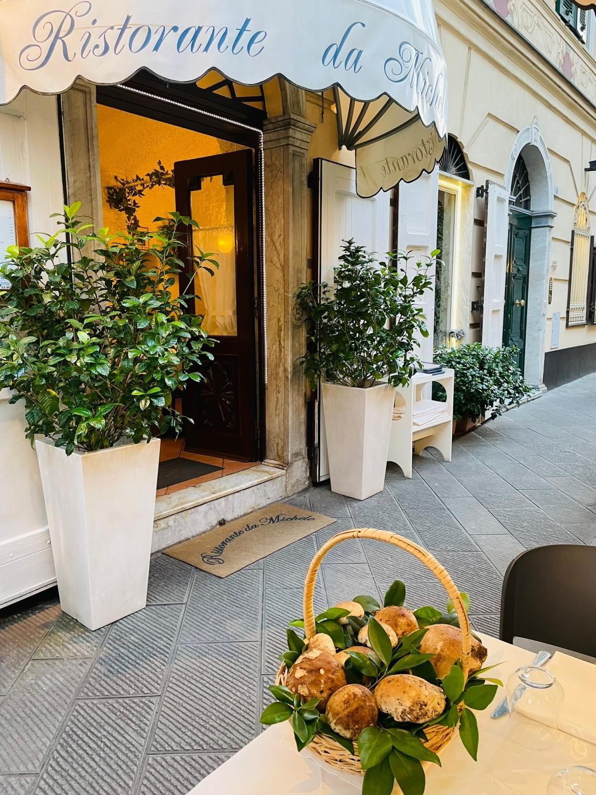 L'ingresso esterno di un ristorante con piante in vaso, una tenda da sole bianca e un cesto di funghi su un tavolo.