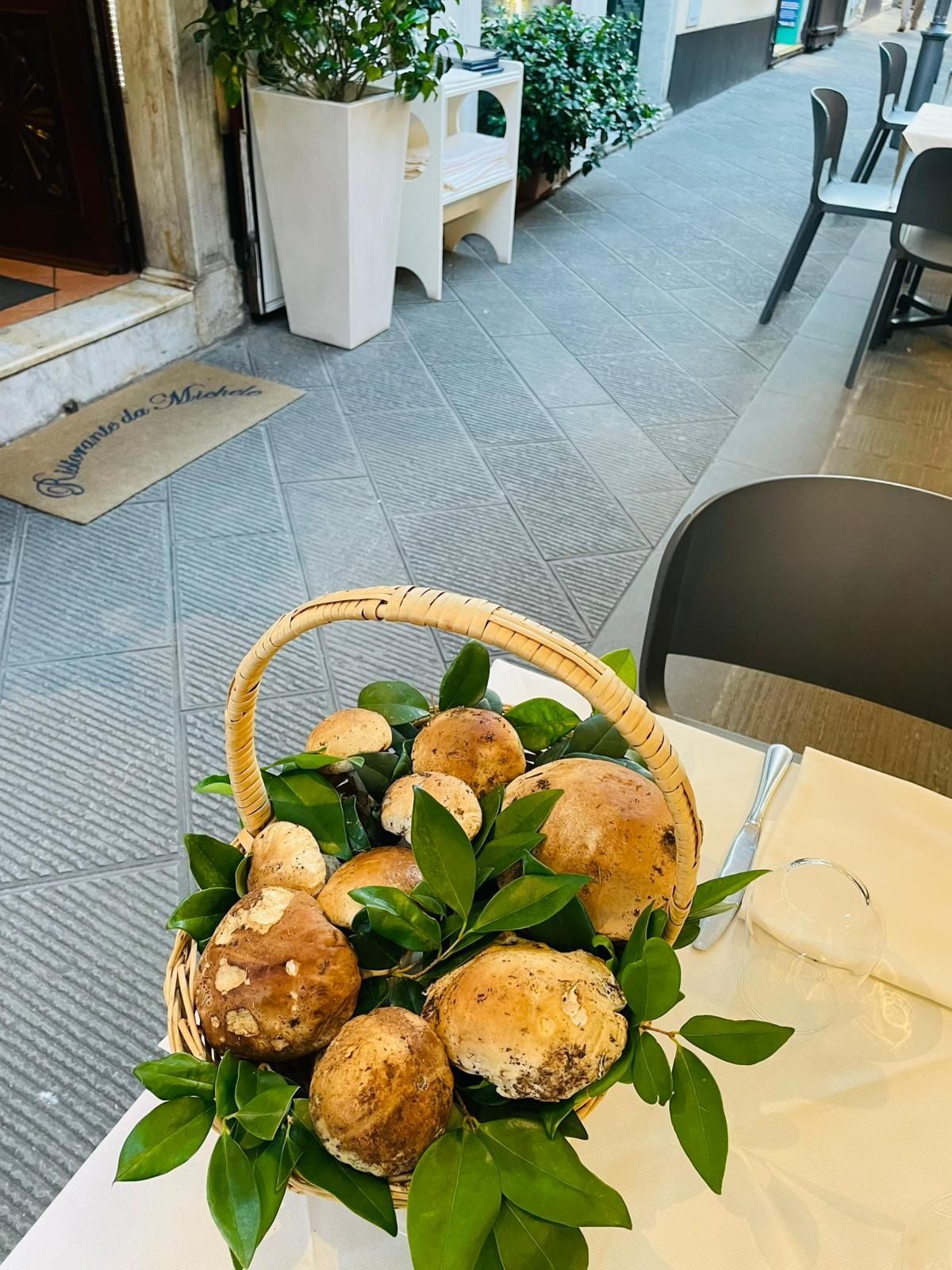 Su un tavolo all'aperto di un ristorante, un cesto di vimini pieno di funghi porcini freschi e foglie verdi è appoggiato.
