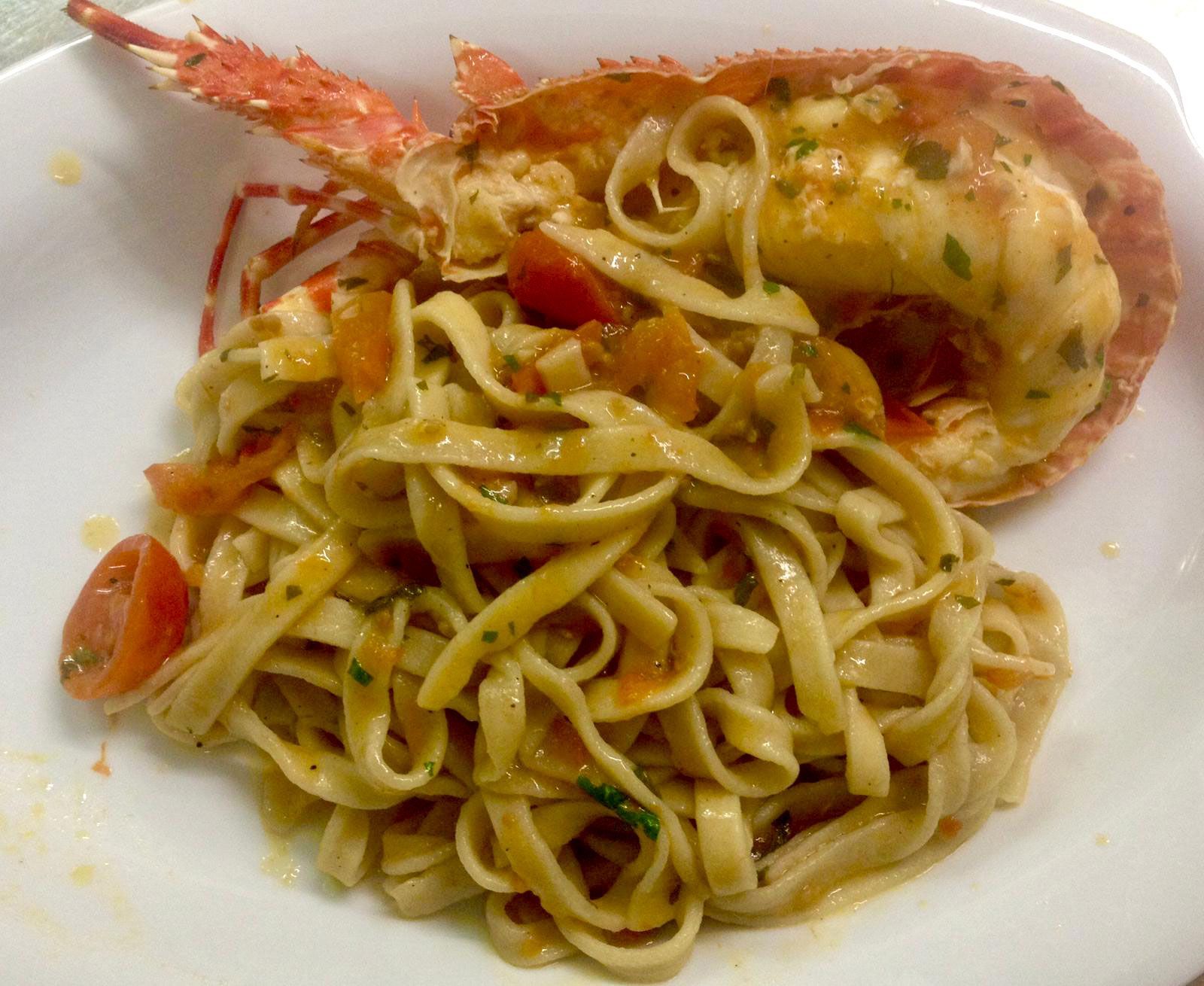 Una ciotola bianca ripiena di linguine, pomodorini e una coda di aragosta grigliata in una salsa leggera.