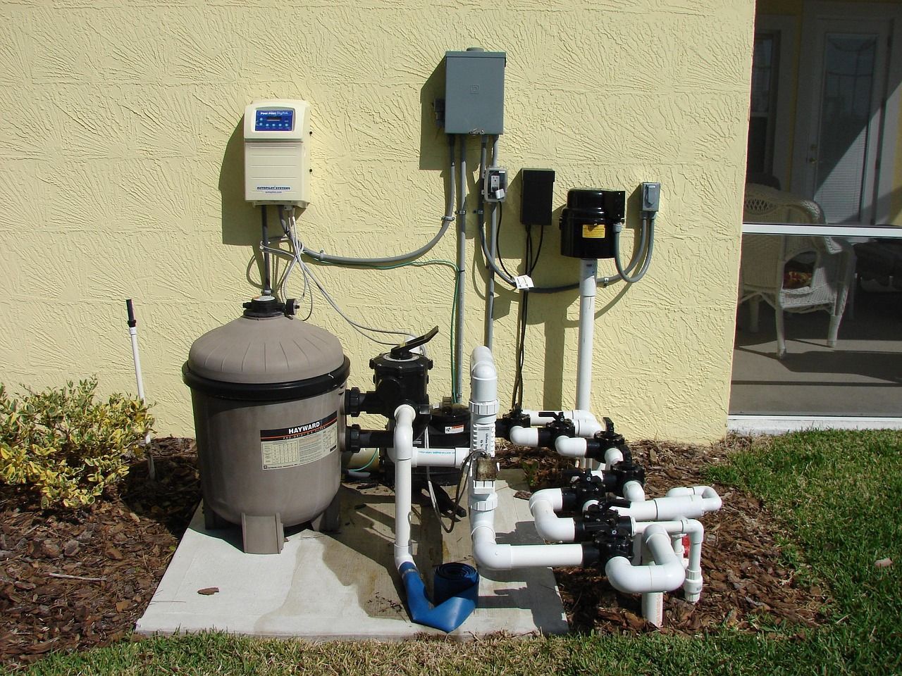 Pool equipment including a filter, control panel, and piping, set against a stucco wall outdoors.