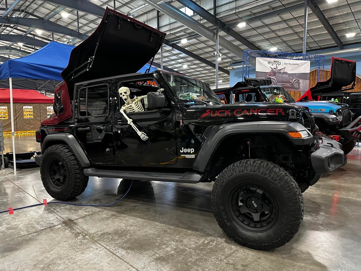 Black off-road Jeep with hood open at an indoor auto show, showing engine and lifted suspension.