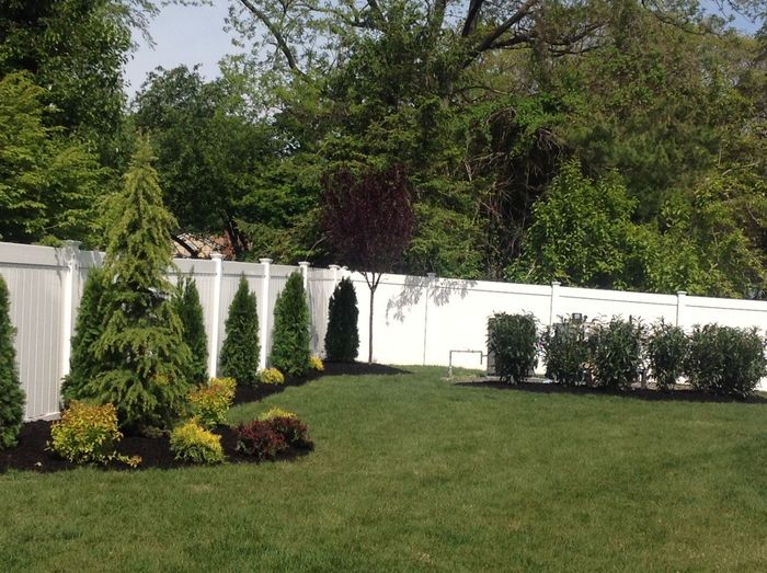 A white fence surrounds a lush green yard with trees and bushes.