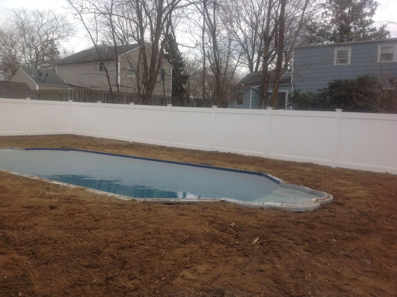 A large swimming pool is surrounded by dirt and a white fence.