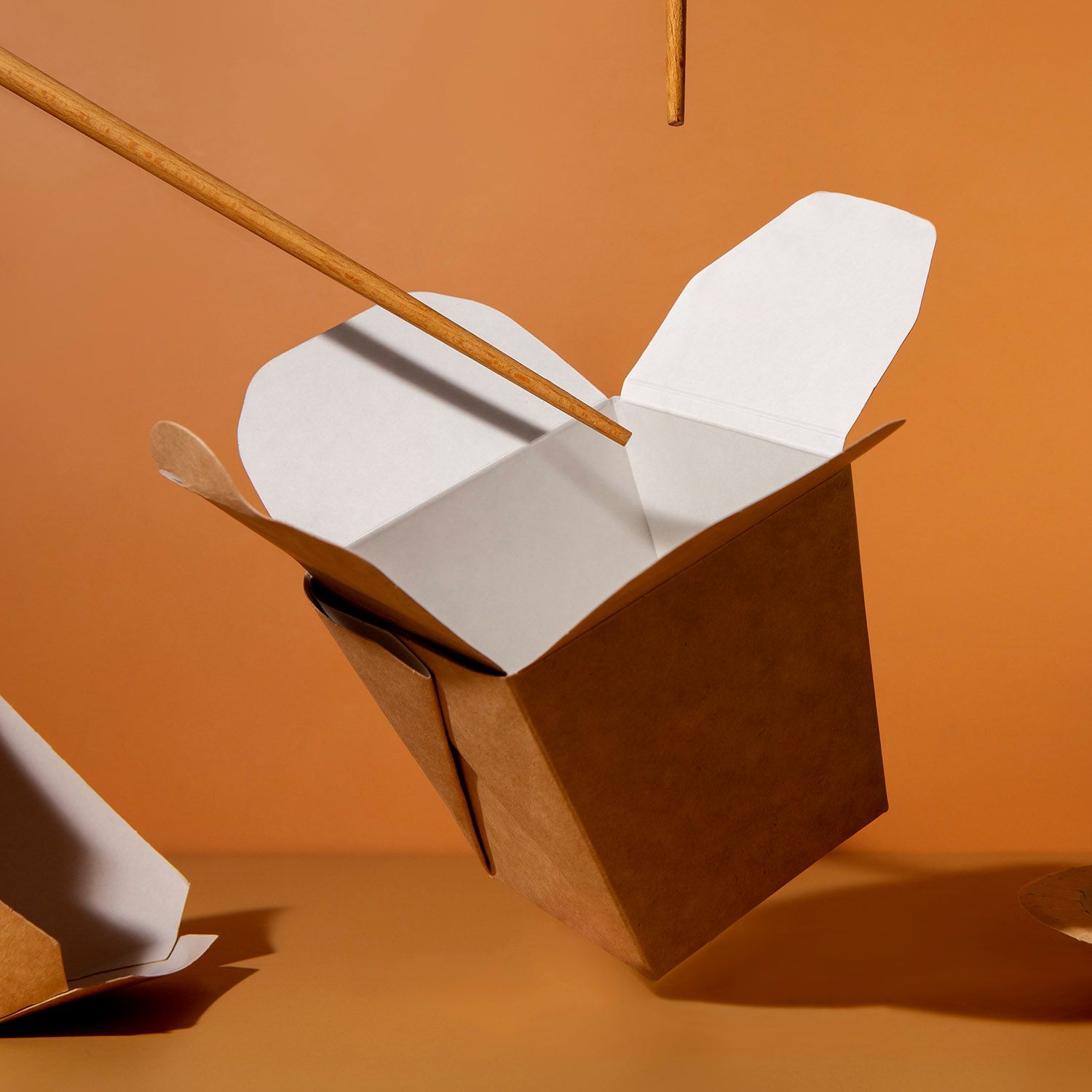 Brown takeout box tipping over, chopsticks reaching in; orange background.