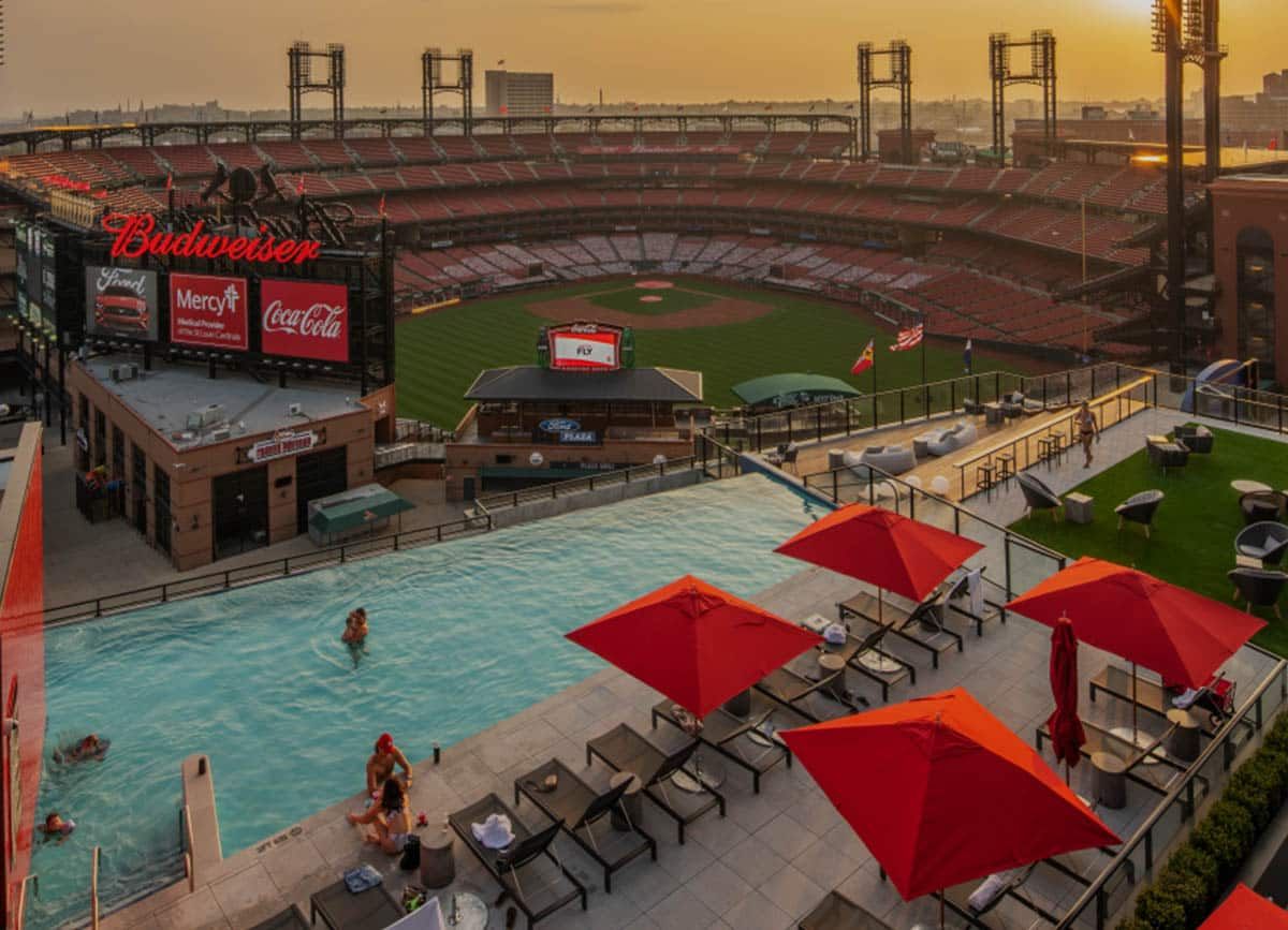 An aerial view of a baseball stadium with a rooftop pool in the foreground, featuring red umbrellas and sunbathers. Sunset casts a warm glow.