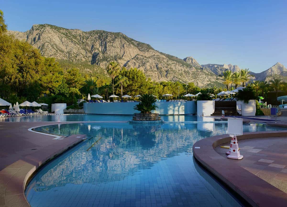 A swimming pool with a mountain backdrop. Clear blue water, blue sky, and trees surround the pool area.