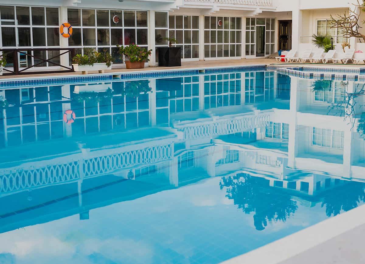 A swimming pool reflects a white building with large windows and a decorative balcony, under a bright sky.