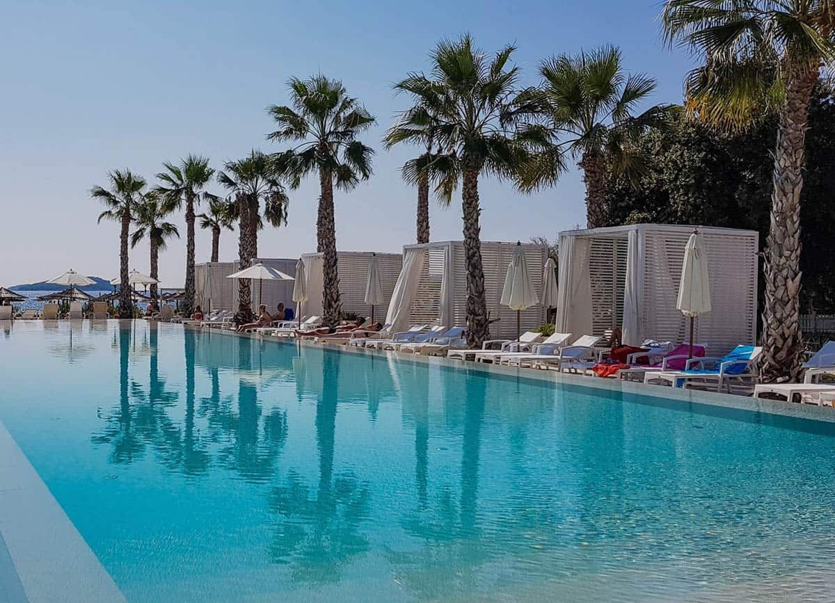 An infinity pool with palm trees reflecting in the clear water. White cabanas and lounge chairs line the pool's edge under a blue sky.