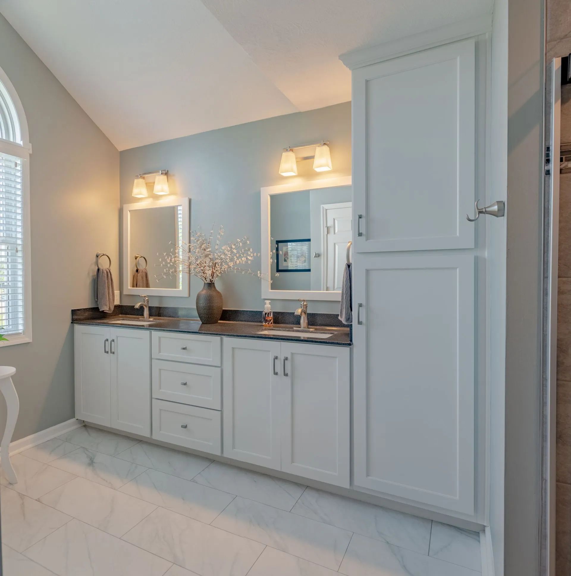 A bright bathroom featuring a white double vanity, two framed mirrors, wall lighting, and grey cabinets on white tile.