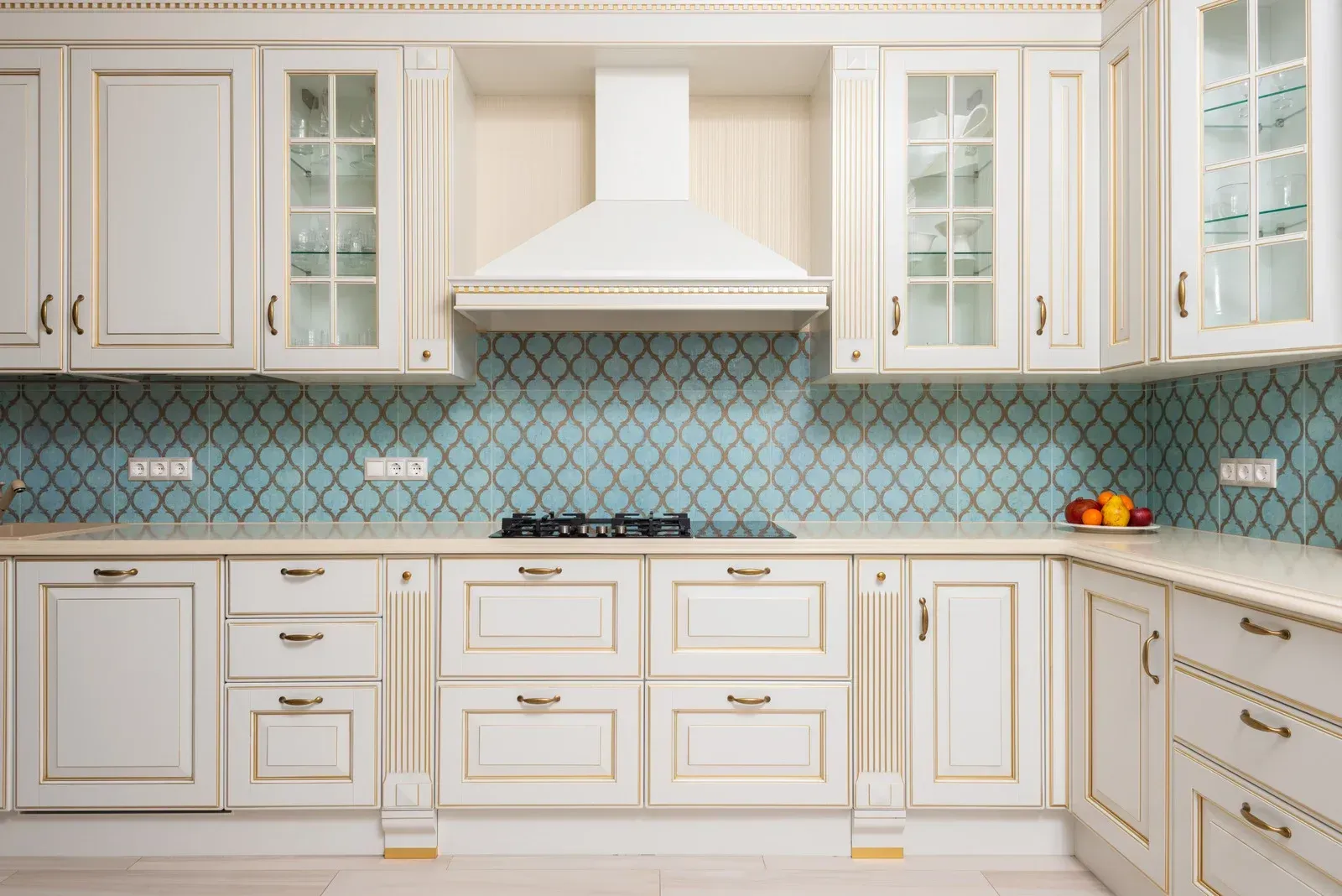 White kitchen cabinets with gold trim, blue patterned backsplash, and built-in range hood.