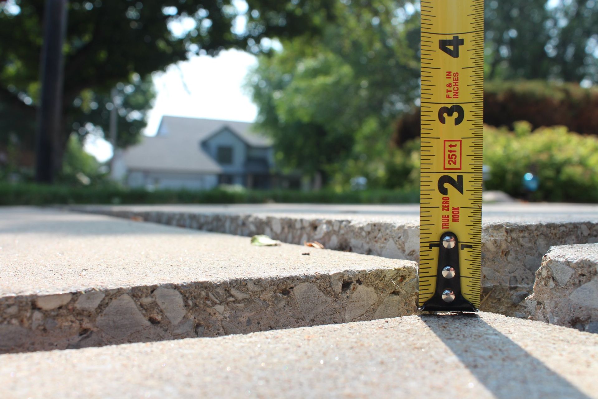Yellow measuring tape on cracked sidewalk; concrete edges with a visible gap; house in background.