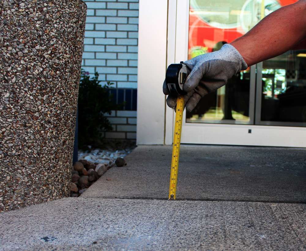 Person in work gloves measuring a concrete sidewalk crack with a yellow tape measure outside a building.