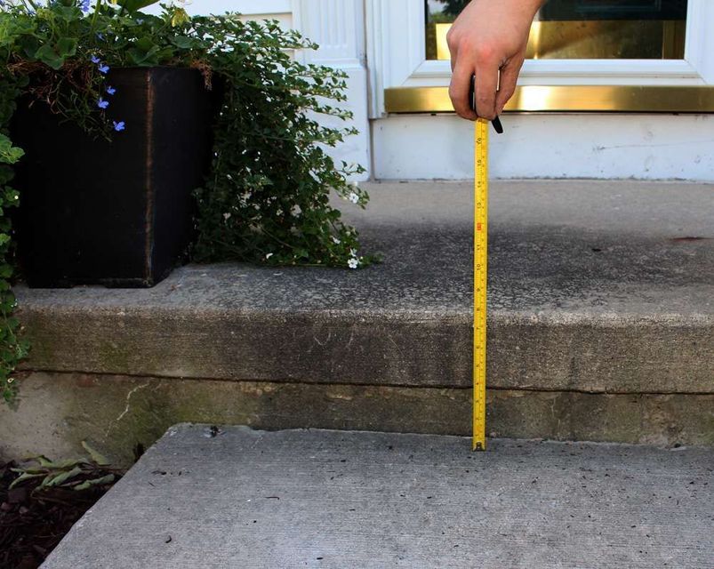 Hand holding a yellow tape measure measuring the height of a concrete step on a porch, next to a black planter.