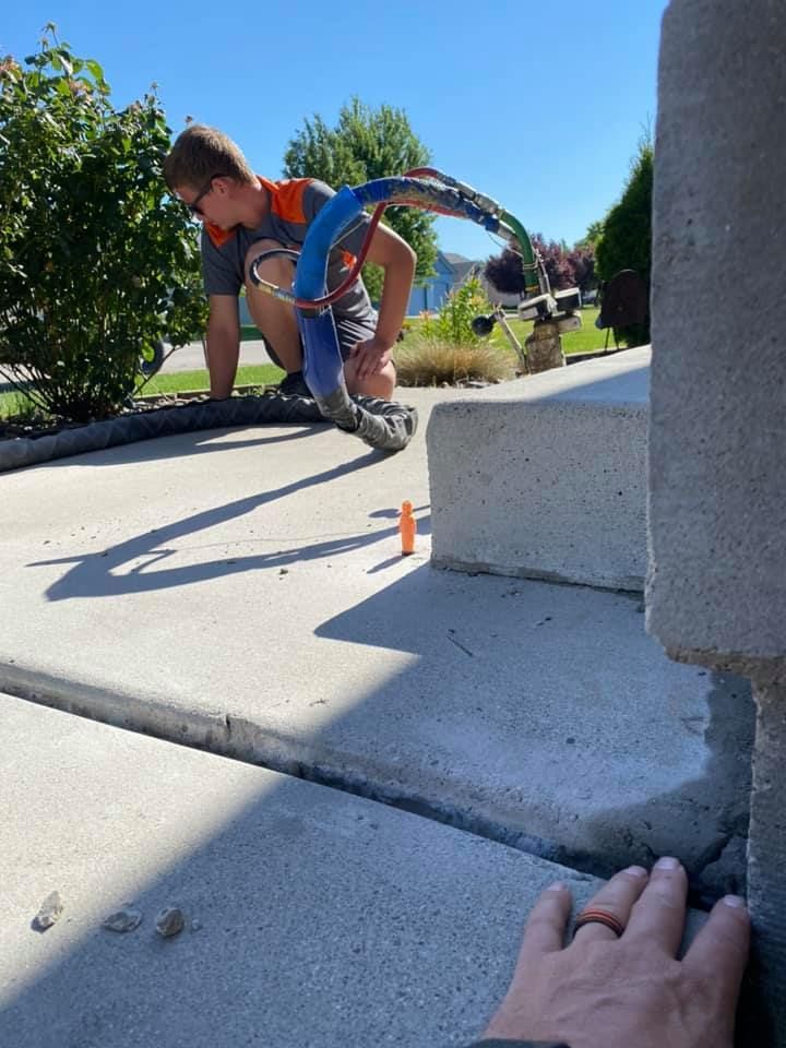 Man kneels near concrete sidewalk crack, holding a device. Sunny outdoor setting.