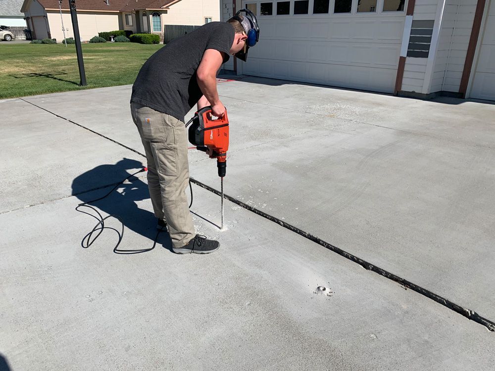 A man in safety glasses uses a jackhammer on a concrete driveway near a garage.