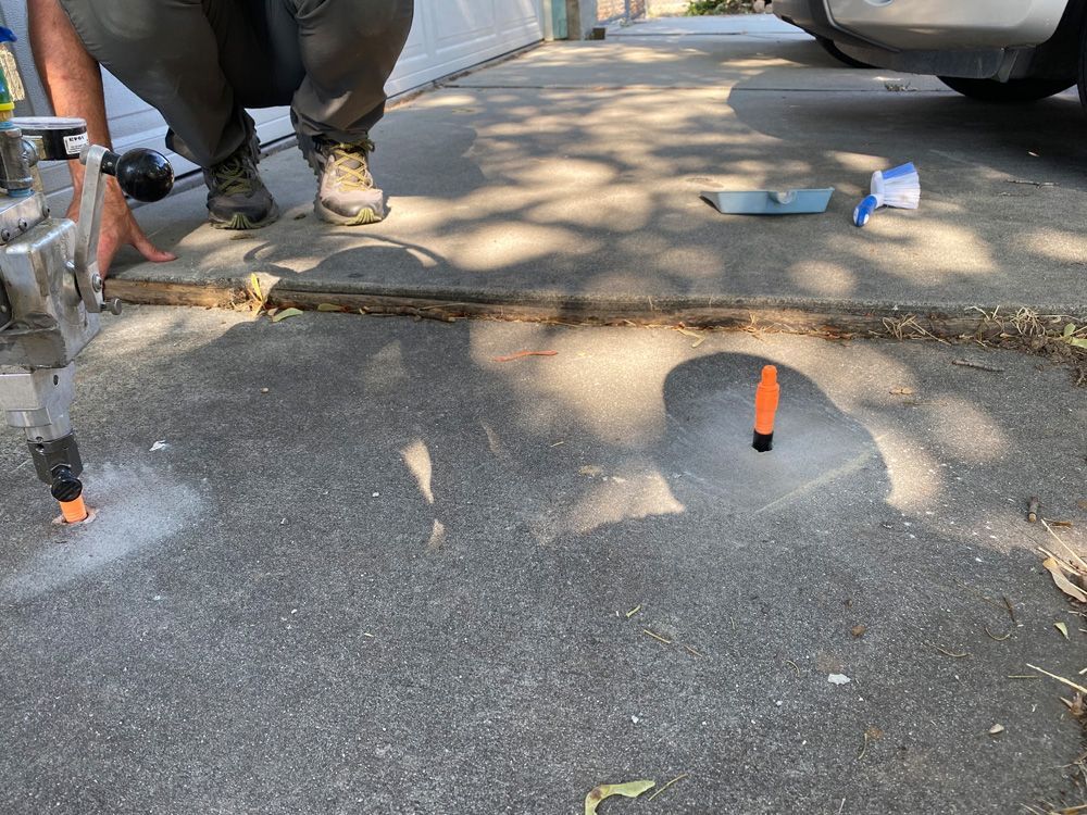 Person using a drill to cut concrete on a driveway; orange markers visible.
