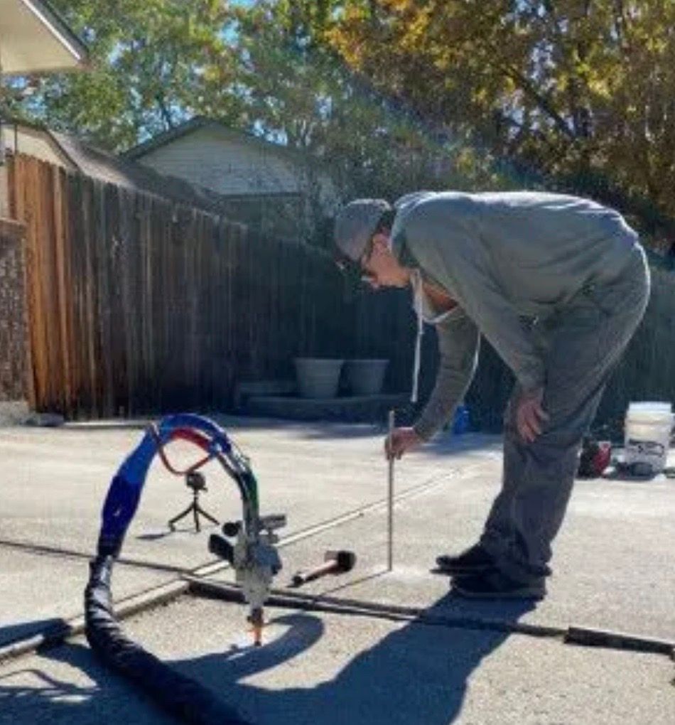 Man using a tool outdoors on concrete. The tool has tubes and nozzles. The setting appears to be a backyard.
