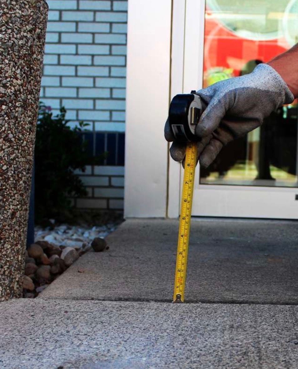 A person with a gloved hand measures the depth of a crack in concrete with a yellow tape measure.