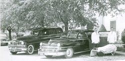 Two black hearses parked in front of a building with two men in white standing beside a covered object.
