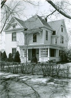 Two-story house with gabled roof, porch, and a dormer. Trees and bushes in the yard.