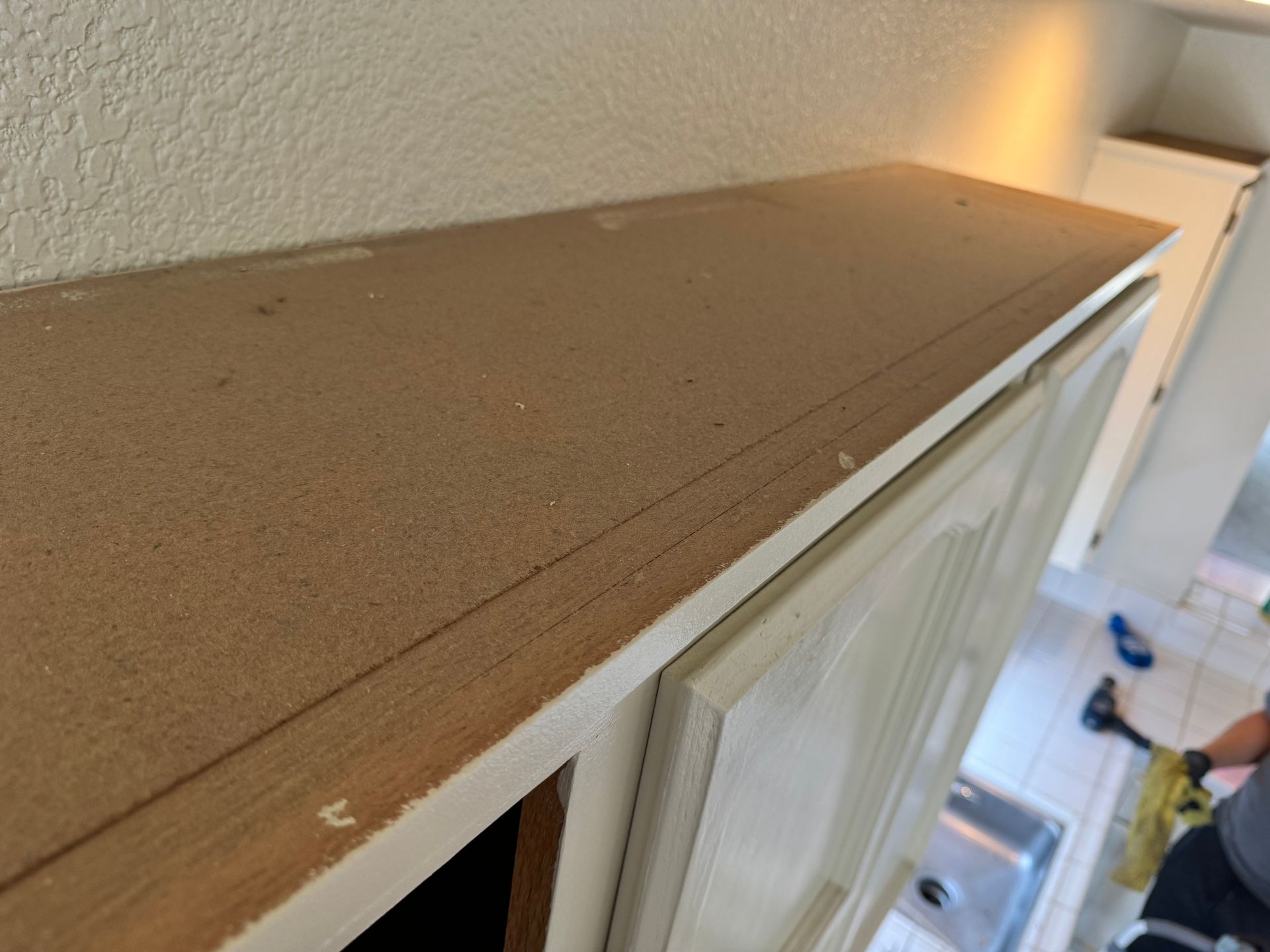 Close-up of the rough, unfinished top surface of white kitchen cabinets, with a blurred view of a sink area below.