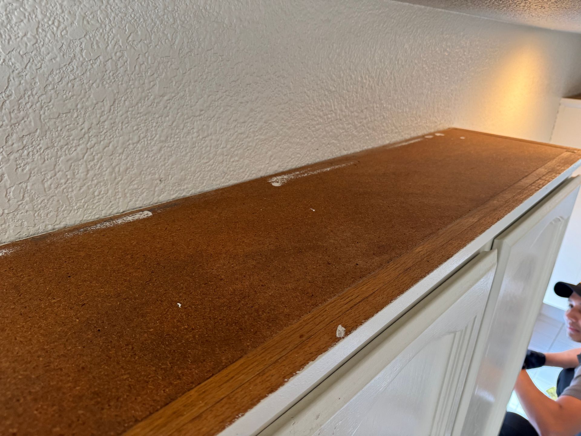 Close-up of a bare particleboard shelf top above white cabinets, with a person working in the blurred background.