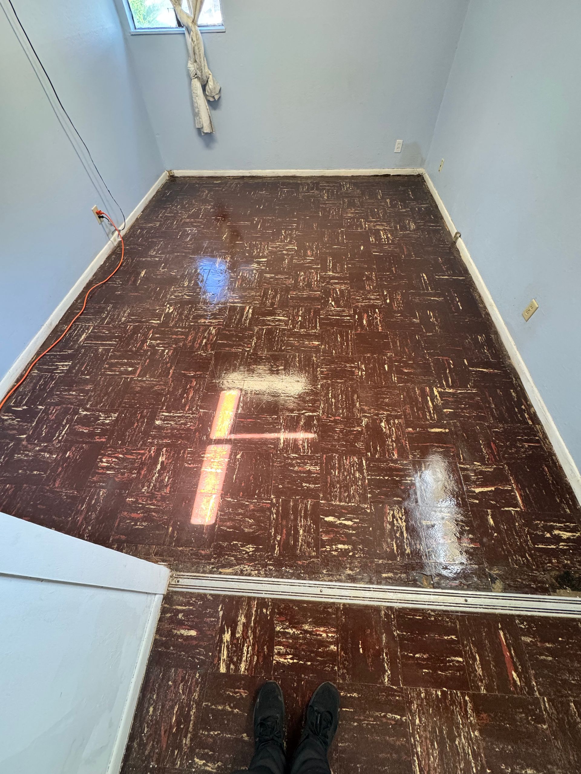 A room with light blue walls and a dark, worn brown patterned floor, with a threshold strip separating two areas.