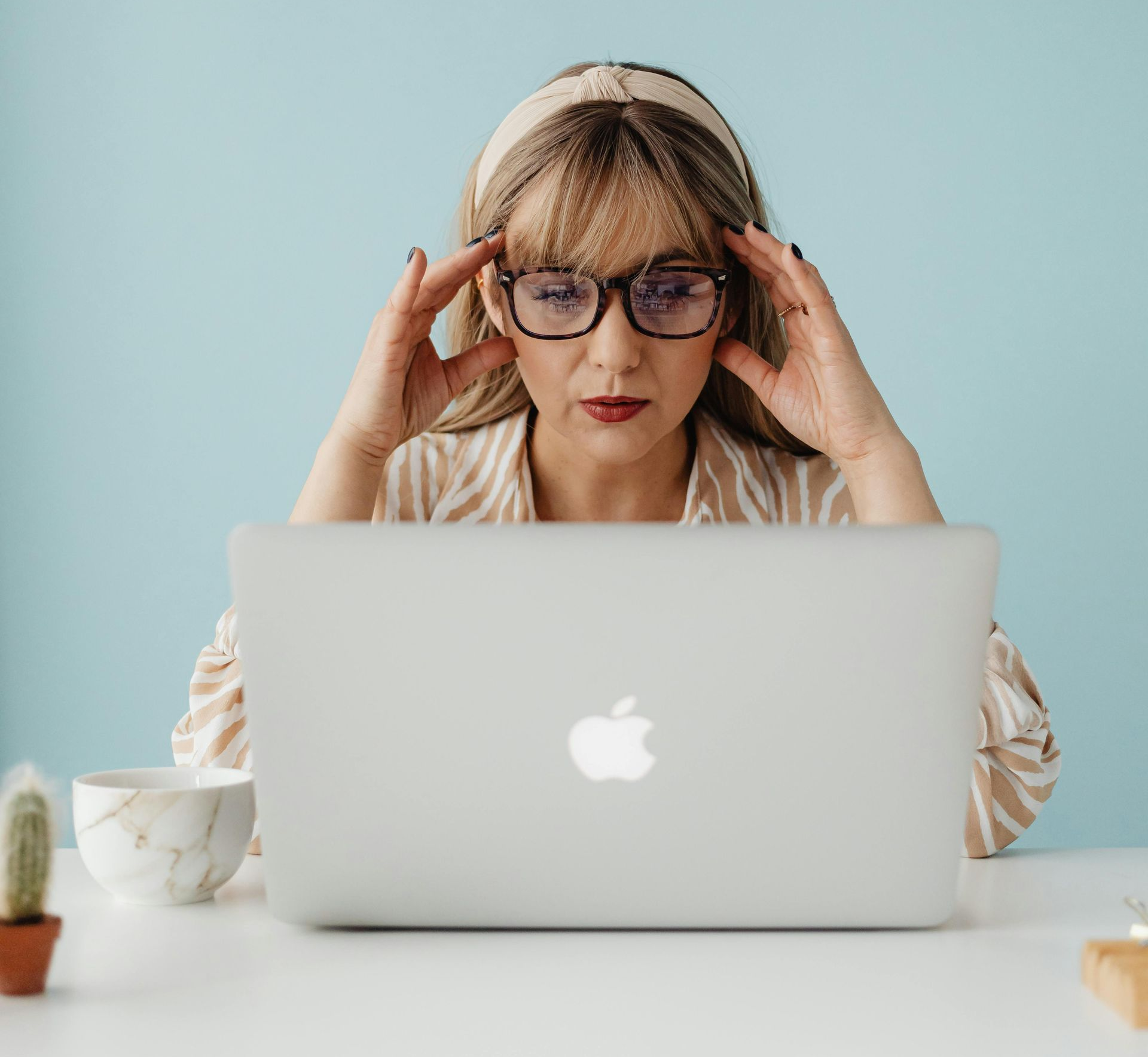 Woman with glasses, hands on head, looking at laptop; light blue background.