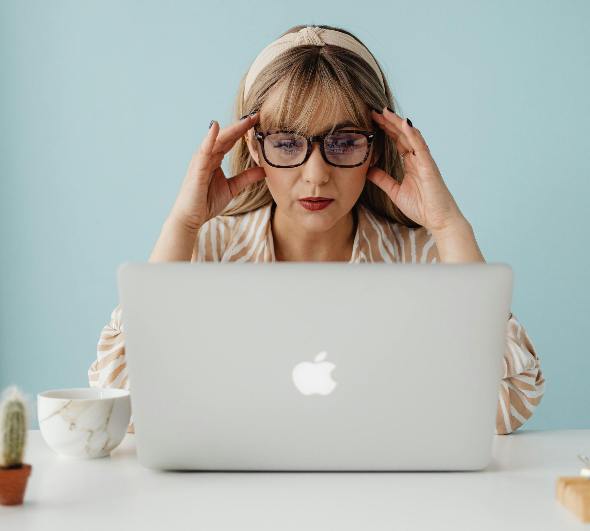Woman at laptop with hands on temples, looking stressed, blue background.