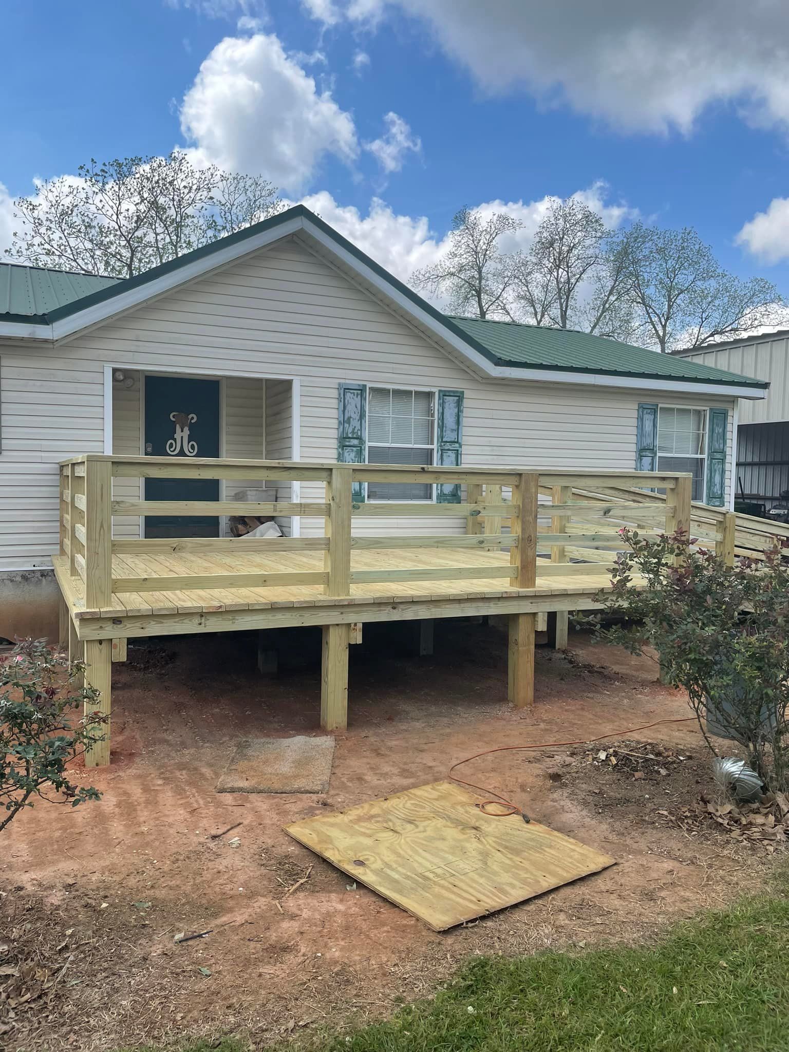 Wooden deck in front of a white house with a green roof. Deck is supported by posts.