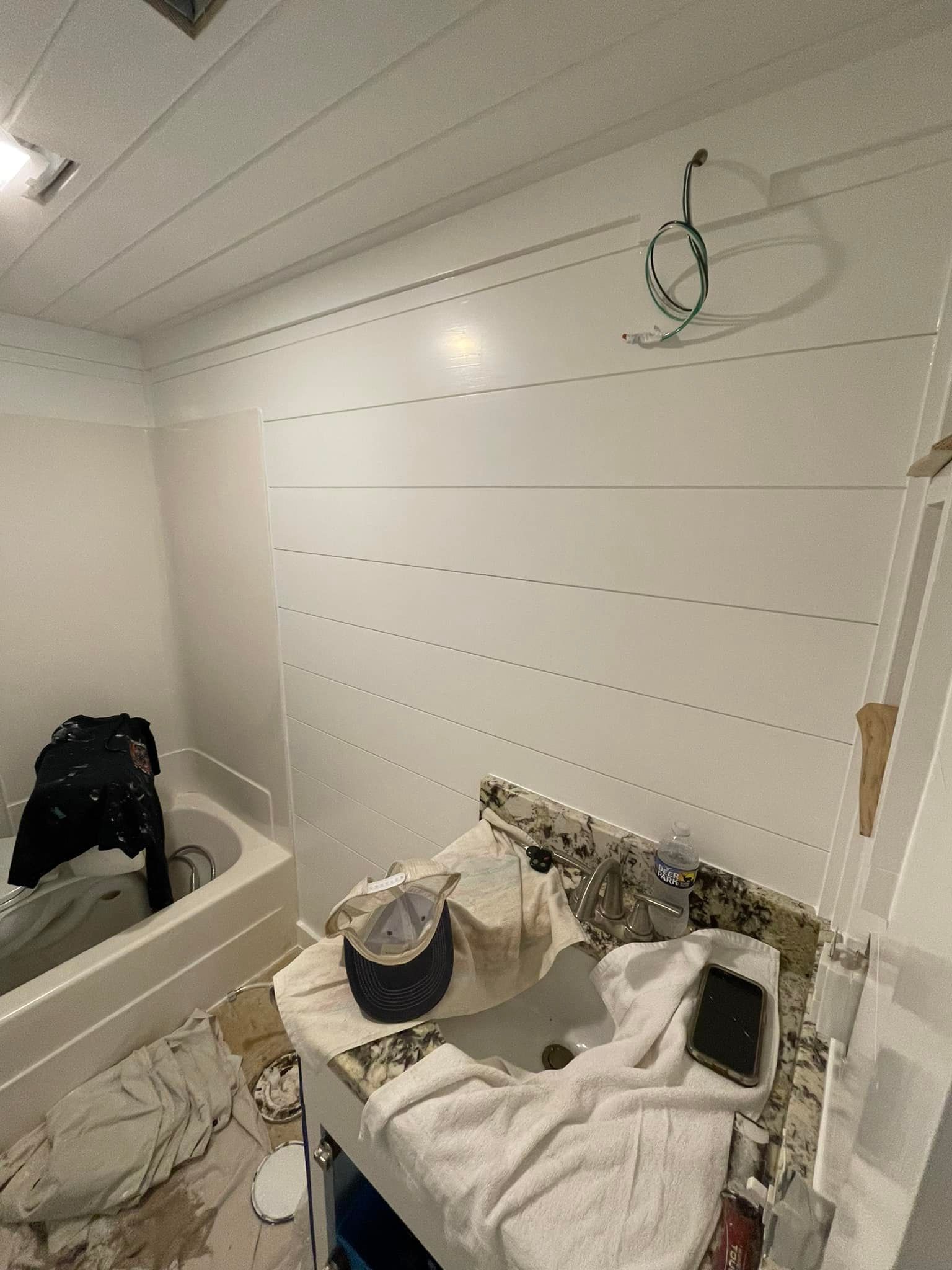 Bathroom with white horizontal wall paneling, vanity, bathtub, and ceiling. Clothes and debris present.