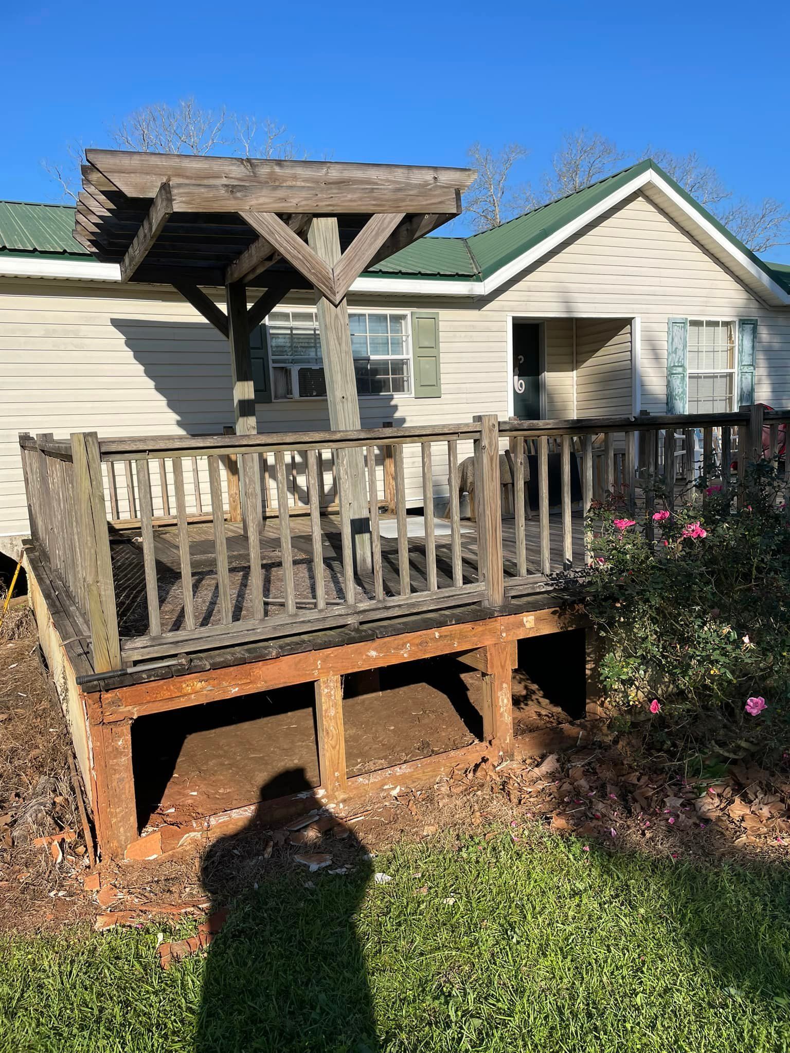 Weathered wooden deck with pergola attached to a light-colored house with a green roof, sunny day.