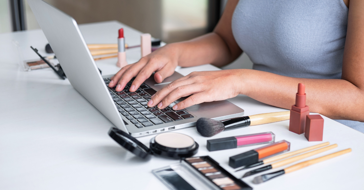 Pessoa analisando tendências de cosméticos no notebook cercada por maquiagens e produtos de beleza