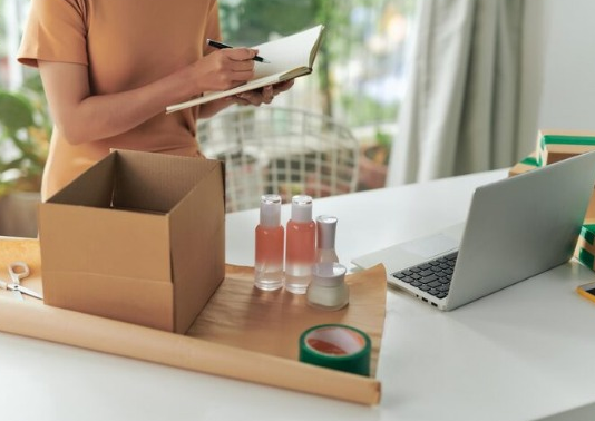 Lojista embalando pedido de cosméticos e usando notebook para controle de estoque no PDV.