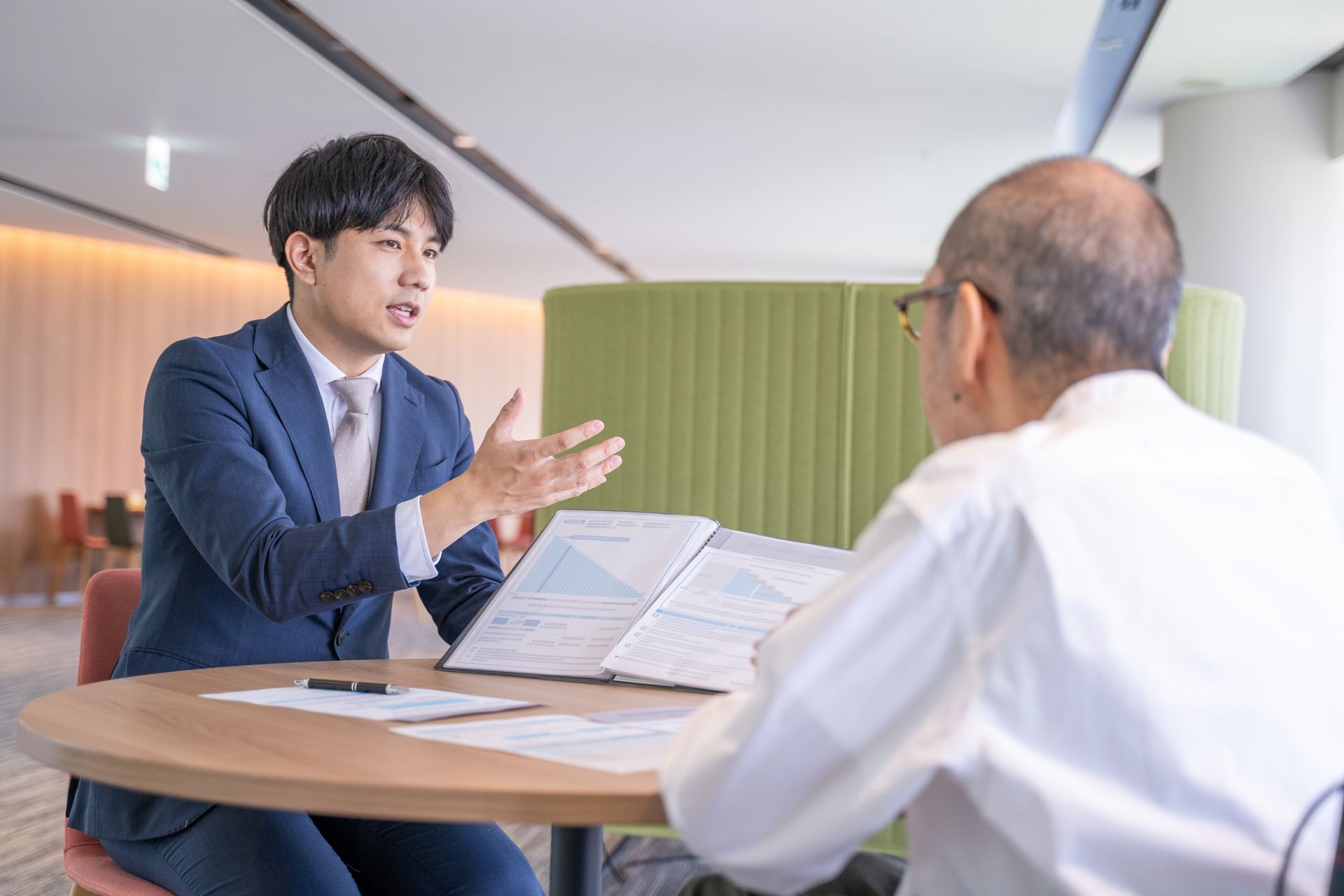Man in suit gesturing, explaining paperwork to older man at a table. Modern office setting.