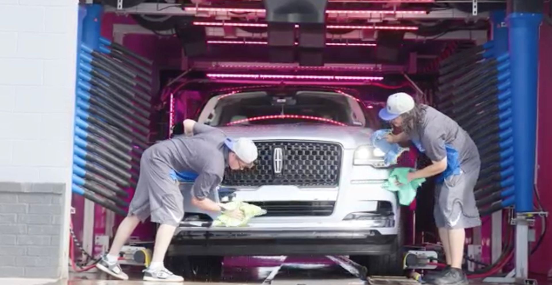 Two workers hand-drying a silver Lincoln in a car wash with pink lighting.