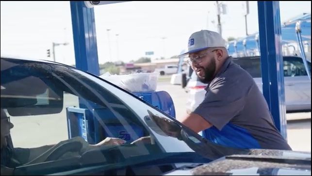 A man in a cap wipes a car's windshield at a gas station.