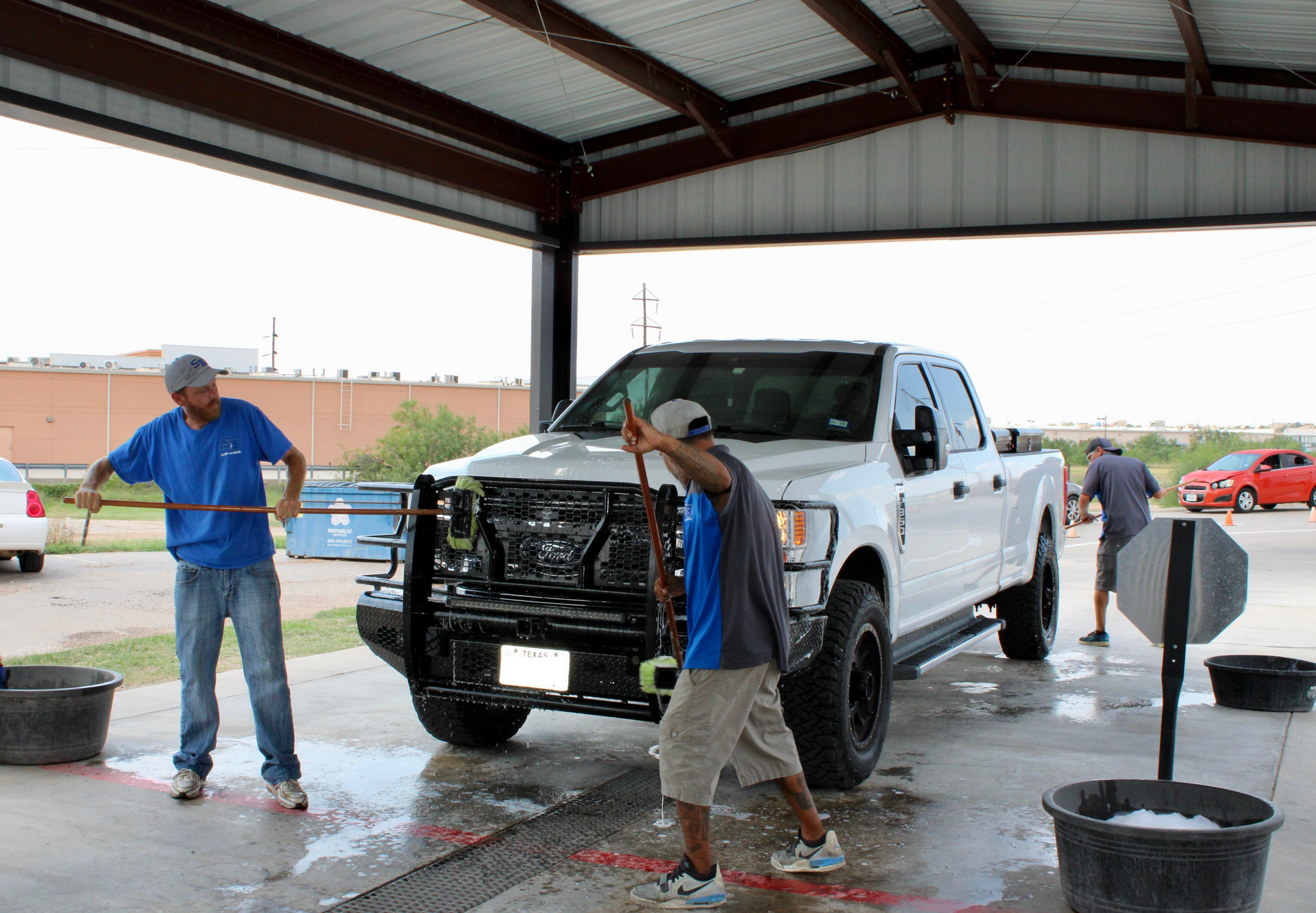 Three people washing a white pickup truck at a car wash; sunny day.