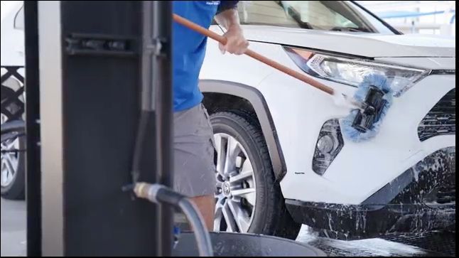 A person washing a white SUV with a long-handled brush at a car wash.