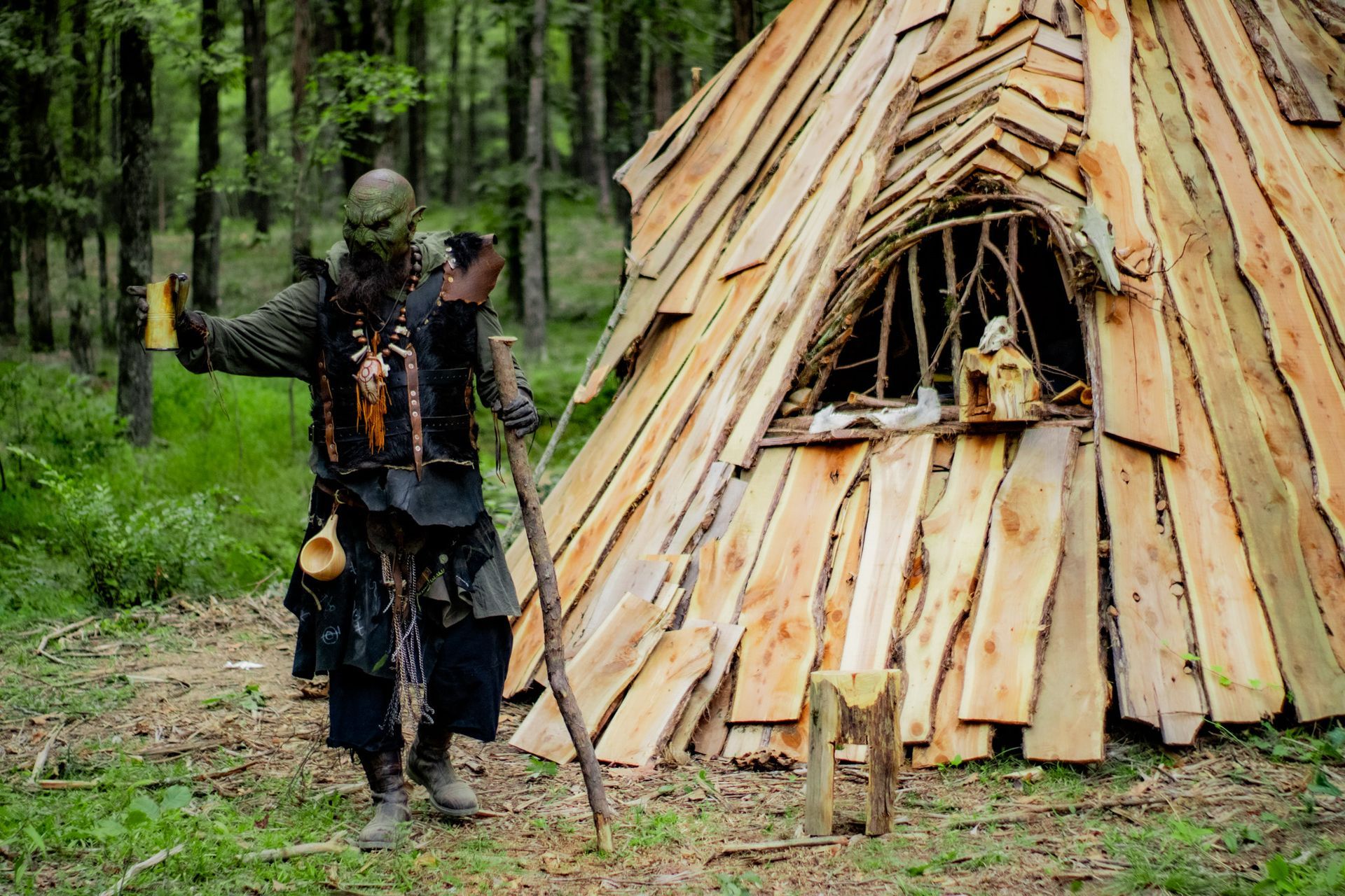 A man in a costume is standing in front of a wooden hut in the woods.