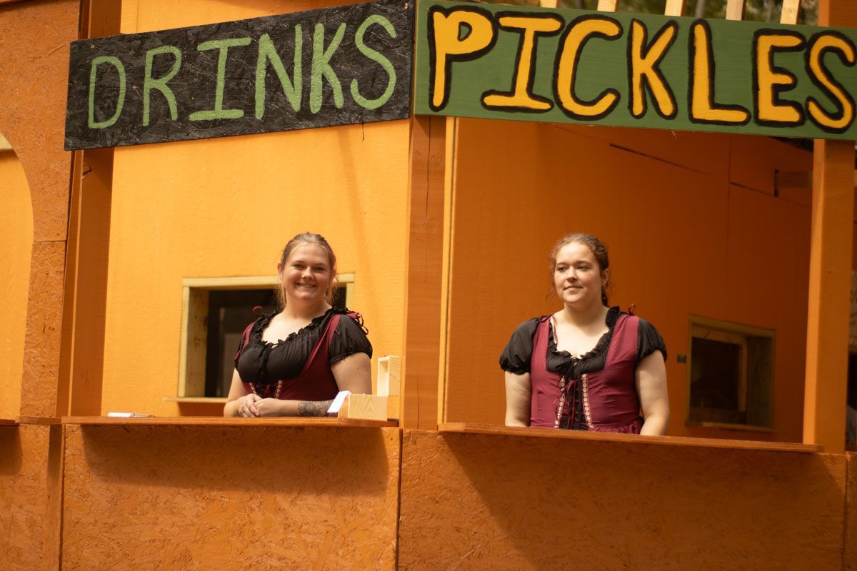 Two women stand behind a counter under a sign that says drinks pickles