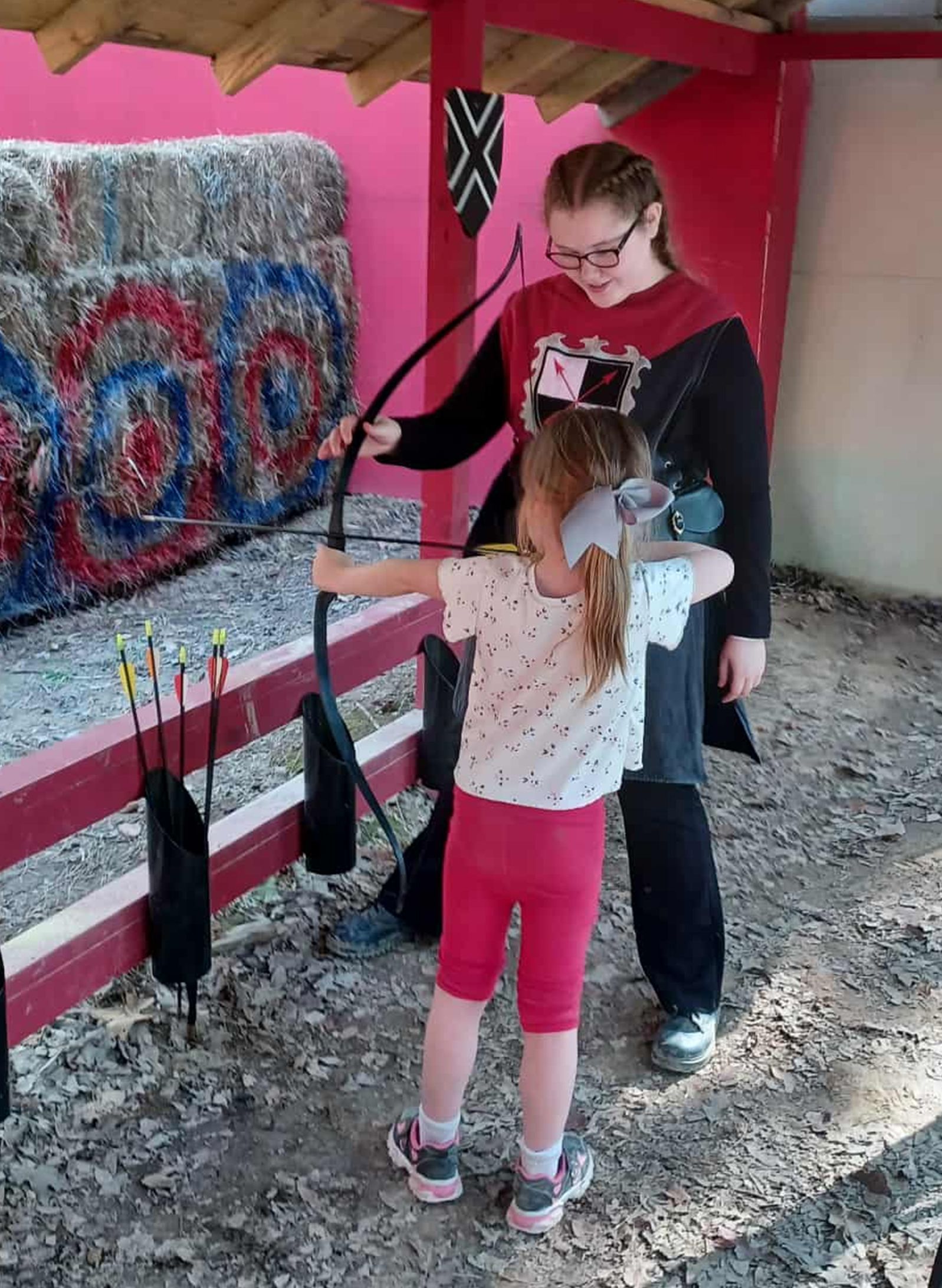 Young person aiming bow and arrow with instructor's help at an archery range.