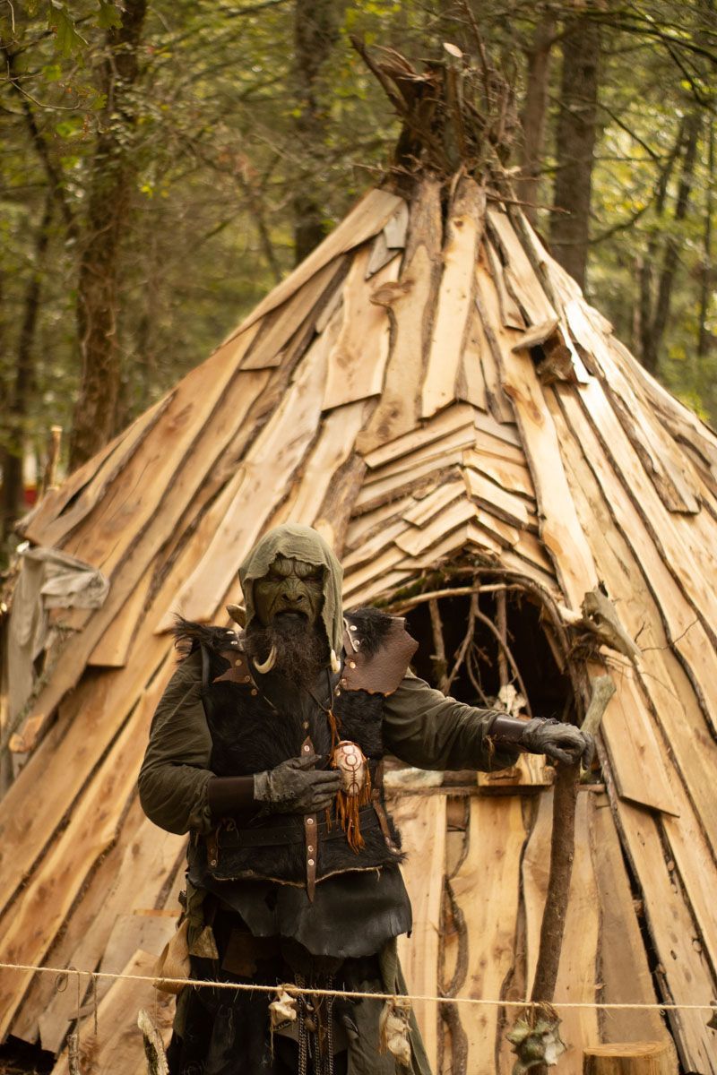 A man in a orc costume is standing in front of a wooden hut in the woods.