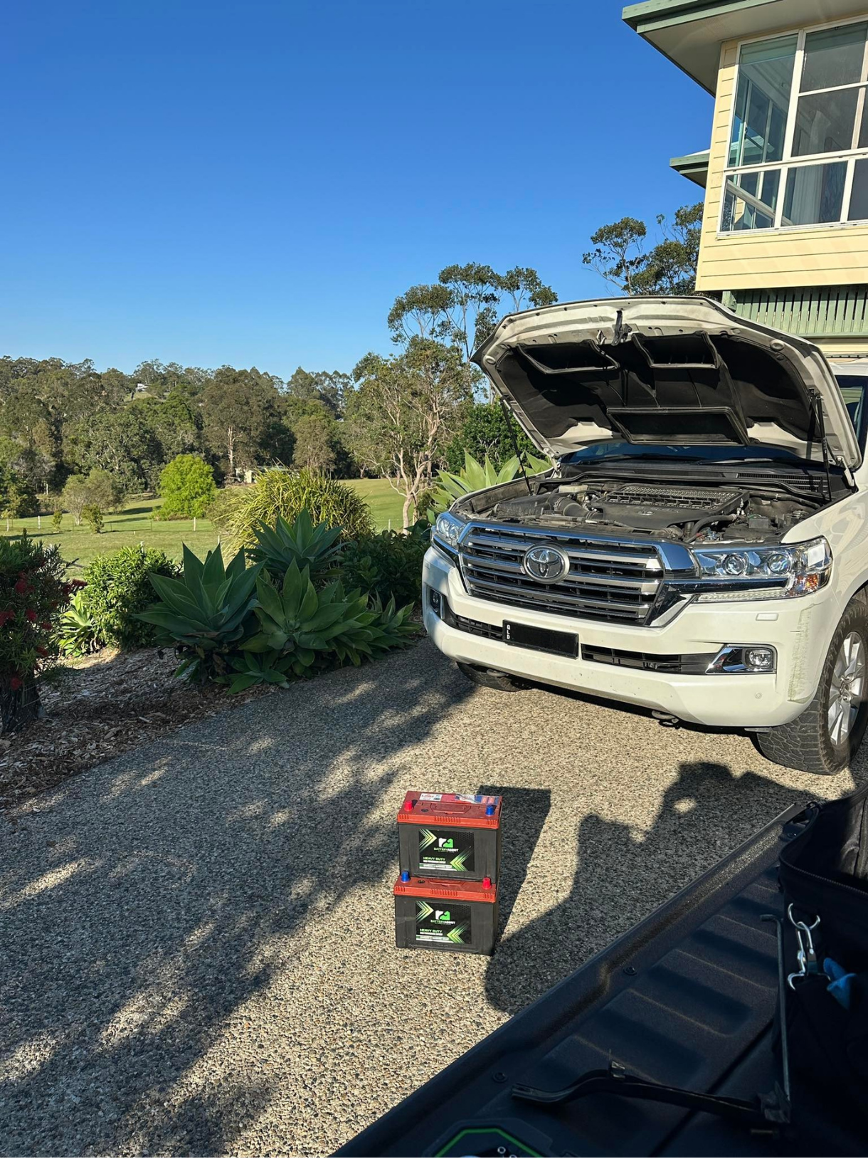 White SUV with open hood and new batteries on a gravel driveway, overlooking a green golf course — Battery Assist QLD In North Lakes, QLD